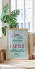 A framed poster with a handwritten message is displayed in a living room, with a potted plant and a bookshelf visible in the background.