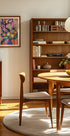 a cozy and well-organized living room with a wooden bookshelf, a round wooden table, and a wooden chair. The room has a warm and inviting atmosphere, with a painting on the wall and a bowl of fruit on the table.
