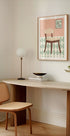 a cozy corner of a room with a wooden desk, a woven chair, a lamp, and a framed artwork depicting a chair on a patterned floor.