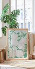 a large framed print of white flowers with green leaves, placed on a wooden floor in a bright, airy room with large windows.